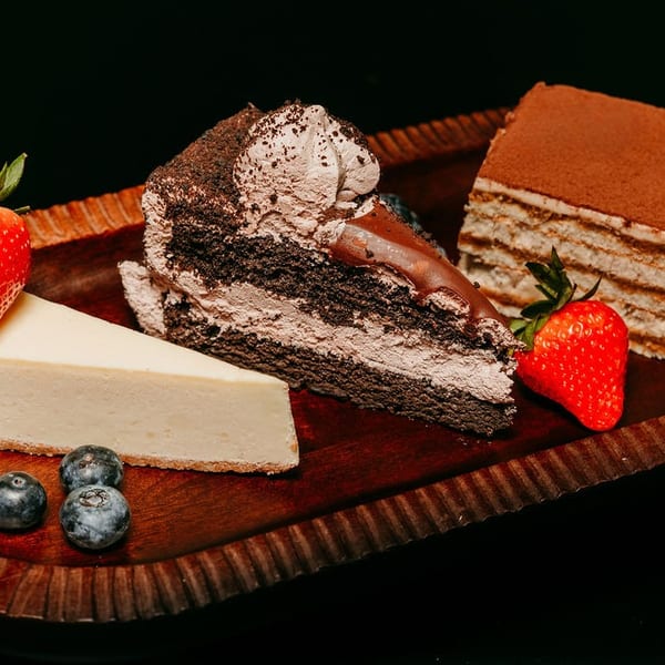 A wooden tray holds slices of cheesecake, chocolate cake, tiramisu, strawberries, blueberries, and blackberries.