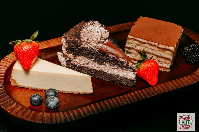 A wooden tray holds slices of cheesecake, chocolate cake, tiramisu, strawberries, blueberries, and blackberries.