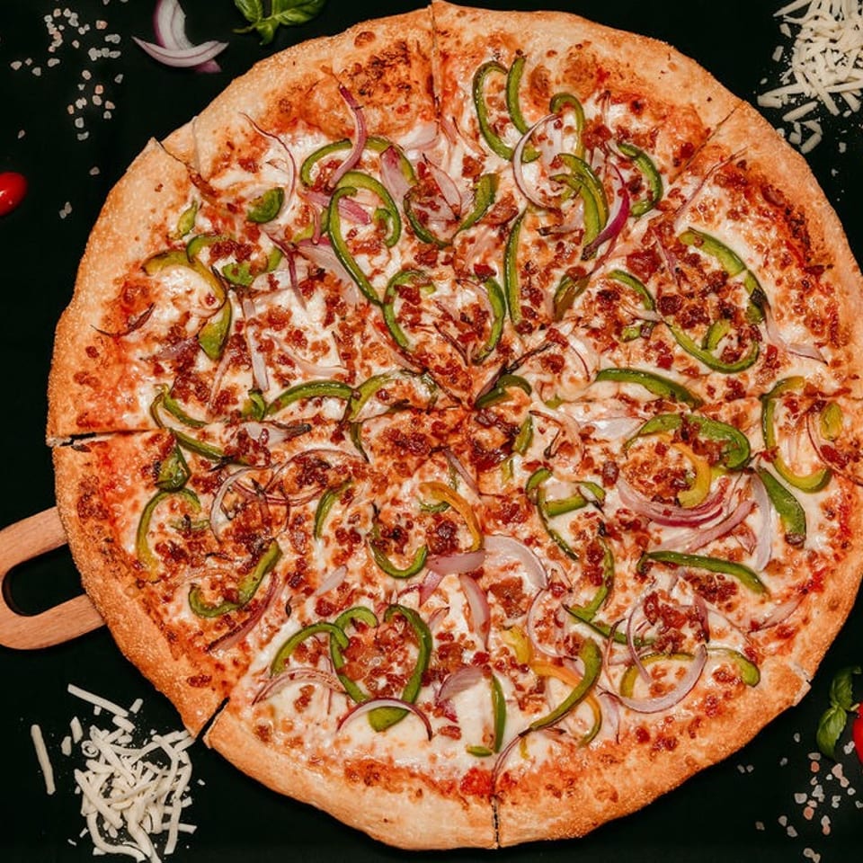 A Fresh Bite Pizza with green peppers, red onions, and bacon bits on a dark surface.