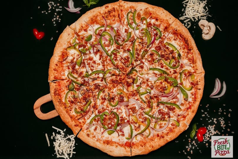 A Fresh Bite Pizza with green peppers, red onions, and bacon bits on a dark surface.