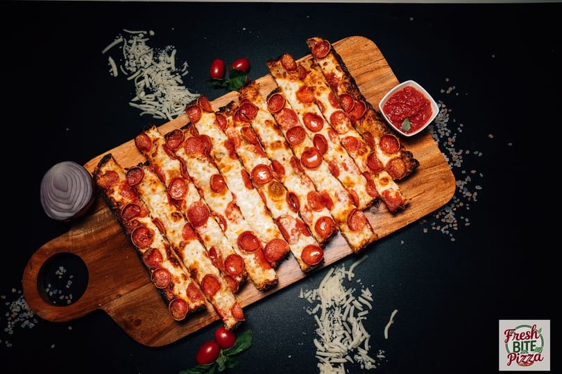Fresh Bite Pizza's pepperoni pizza strips with dipping sauce, cheese, and tomatoes on a wooden board.