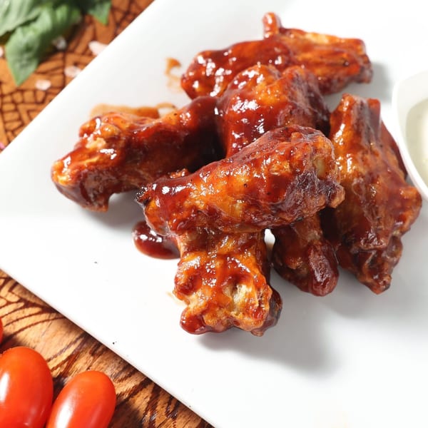 Glazed chicken wings on a white plate with a side of dipping sauce, basil, and cherry tomatoes.