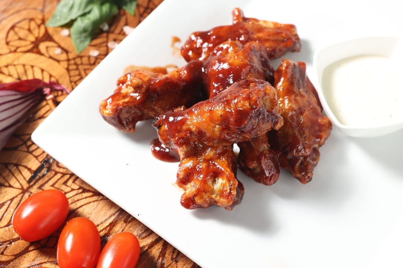 Glazed chicken wings on a white plate with a side of dipping sauce, basil, and cherry tomatoes.
