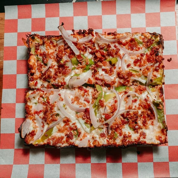 Rectangular pizza with bacon, green peppers, and onions on checkered paper and a wooden board.