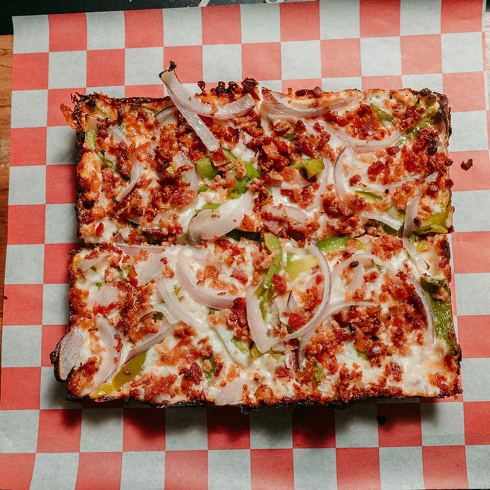 Rectangular pizza with bacon, green peppers, and onions on checkered paper and a wooden board.