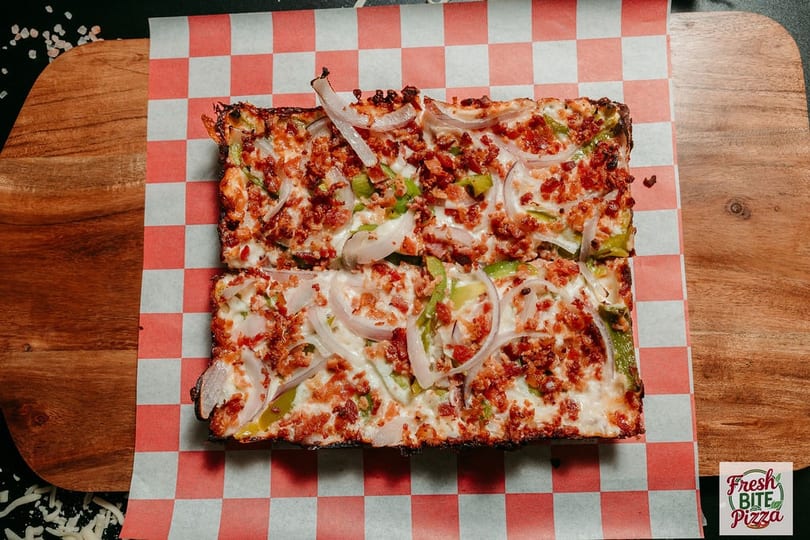 Rectangular pizza with bacon, green peppers, and onions on checkered paper and a wooden board.